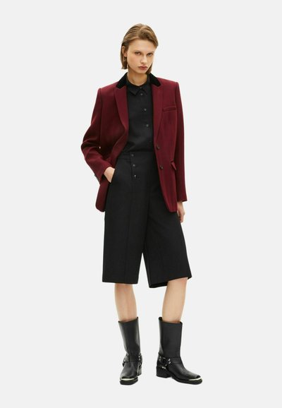 Blazer bordeaux con colletto nero, camicia nera, pantaloni corti larghi neri e stivaletti neri alla caviglia. Presenta bottoni e un dettaglio tasca sul blazer.