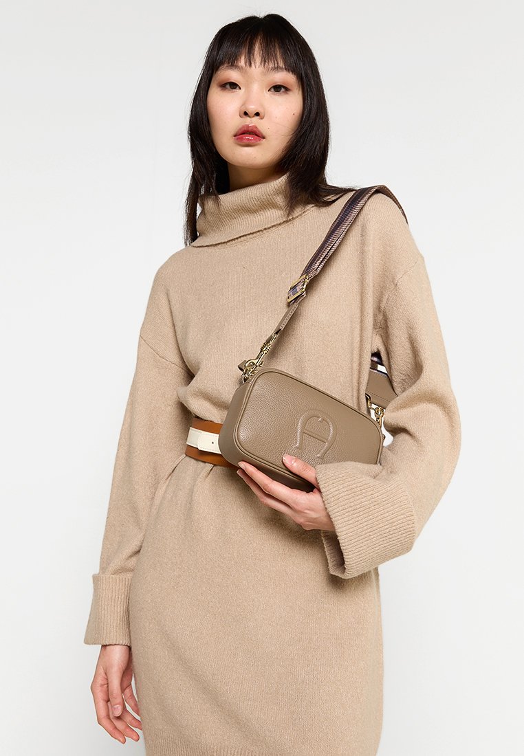 Braune Leder-Crossbody-Tasche mit geprägt Logo, mit strukturierter Oberfläche und einem gemusterten verstellbaren Trageriemen.getragen über einem cremefarbenen Strickkleid.