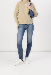 Beige ribbed sweater with a round neck and logo patch, paired with blue skinny jeans and white sneakers featuring black accents.