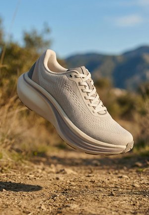 Bež tekaški čevelj z oblazinjeno podplato, ki plava nad peščeno poti, z zamegljeno vegetacijo in gorami v ozadju.