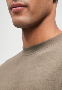 Καφέ βαμβακερό πουλόβερ με στρογγυλή λαιμόκοψη και λεία υφή, ριγέ γιακά και διακριτές ραφές, εμφανισμένο γύρω από τη λαιμόκοψη και τη γραμμή της σιαγόνας.