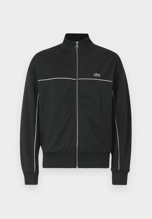 Giacca nera con zip e colletto alto, piping bianco sul petto e logo Lacoste verde sul lato sinistro. Polsi e orlo a coste.