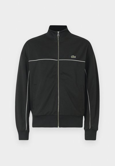 Veste zippée noire avec un col haut, un piping blanc sur la poitrine et un logo Lacoste vert sur le côté gauche. Poignets et ourlet côtelés.