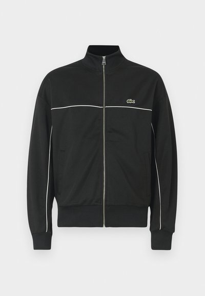 Veste zippée noire avec un col haut, un piping blanc sur la poitrine et un logo Lacoste vert sur le côté gauche. Poignets et ourlet côtelés.