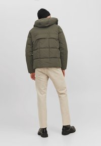 Giubbotto imbottito verde con cappuccio, caratterizzato da un design trapuntato orizzontale. Indossato con pantaloni beige e stivali neri. Tessuto liscio.