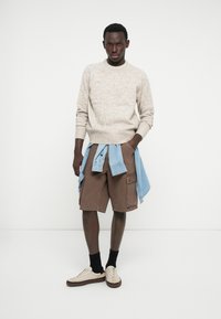 Beiger Pullover mit strukturierter Strickmuster, braune Cargoshorts und ein hellblaues Hemd, um die Taille gebunden. Paar cremefarbene Sneakers mit dunklen Sohlen.