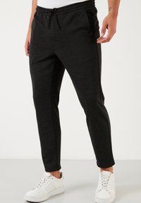 Černé teplákové kalhoty s texturou, stahovací šňůrkou v pase a bočními kapsami, úzký střih zúžený k kotníkům, v kombinaci s bílými teniskami.