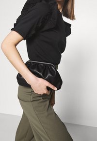 Kvinna som håller en liten svart rynkad clutchväska, iklädd en svart topp med volanger och olivgröna byxor mot en enfärgad bakgrund.