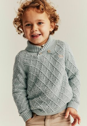 Krøllet-håret småbarn smilende iført en tekstureret lyseblå strikket sweater med knapper og beige bukser, stående mod en ensfarvet baggrund.