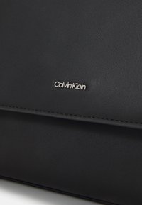 Black textured leather surface with stitched edge and small silver Calvin Klein logo centered above the seam.