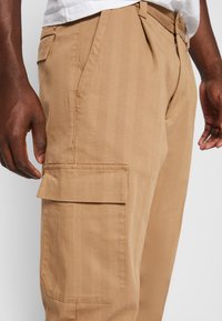 Cargo-Shorts aus Baumwolle in Beige mit Seitentasche, geradem Schnitt und sichtbaren Nähten. Der Stoff hat eine dezente Textur und feine vertikale Streifen.
