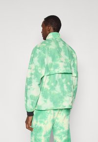 Πράσινο μπουφάν με το μοτίβο tie-dye, ψηλό γιακά, χαλαρή εφαρμογή και ελαστικά μανίκια. Το ύφασμα έχει μαλακή υφή και δίχρωμο σχέδιο, συνδυασμένο με παντελόνια στο ίδιο χρώμα.