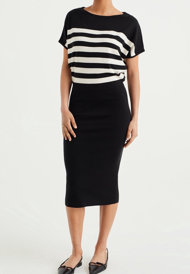 Femme portant un haut à manches courtes à rayures horizontales noires et blanches, une jupe crayon noire longueur genou et des ballerines noires pointues.