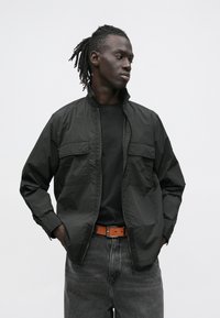Black lightweight jacket with stand collar, two chest pockets, zip closure, worn over a black shirt and gray denim, accented by a brown belt.