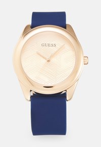 Montre dorée avec un cadran rose clair à motif, bracelet en silicone bleu, boîtier rond et une couronne sur le côté. Marque visible : "GUESS."
