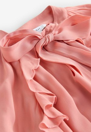 Pink chiffon bluse med en knyttet sløjfe ved halsen, med bølgede kanter og blød tekstur, der fremhæver det flydende stofdesign.