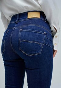 Jeans de mezclilla azul oscuro con un diseño ajustado, que presentan un parche trasero de cuero marrón, bolsillos visibles y costuras en contraste.