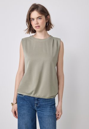 Jeune femme aux cheveux bruns courts portant un haut sans manches beige, un jean bleu, des boucles d'oreilles créoles et un bracelet en or, sur fond uni.