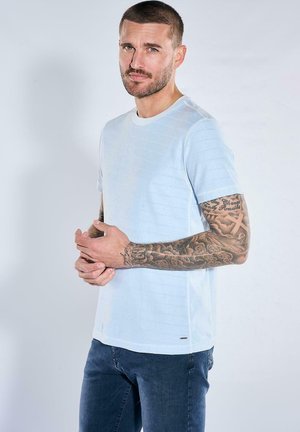 Mann mit kurzem Haar und Bart, der ein hellblaues kurzärmliges Hemd und Jeans trägt und tätowierte Unterarme zeigt, vor einem schlichten Hintergrund.