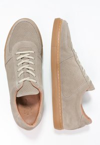 Beige mockasin sneakers med perforerad detalj och beige snören, med en brun gummisula och kontrasterande detaljer runt öppningen.