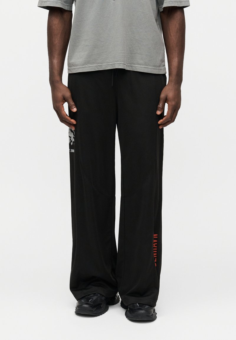 Čierne športové nohavice s voľným strihom, hladkou textúrou a detailom loga na boku. Kombinované so sivým tričkom a čiernou obuvou.