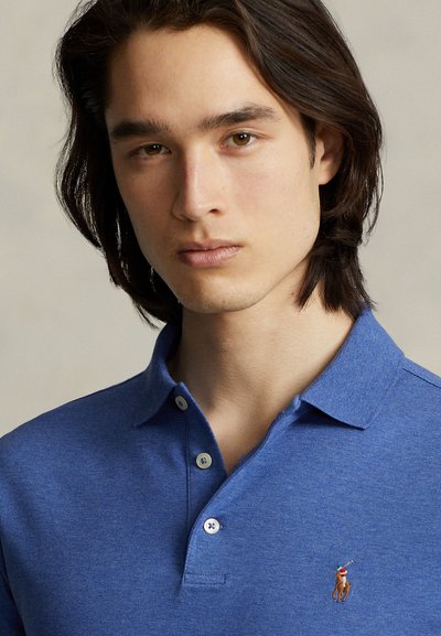 Fiatal férfi középhosszú sötét hajjal, kék galléros pólót visel, amelyen egy kis hímzett ló és lovas logó található.