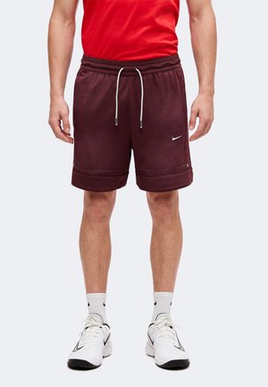 Homme portant un short Nike bordeaux avec un cordon, des chaussettes blanches Nike et des baskets blanches Nike, debout devant un fond uni.