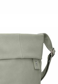 Bolso cruzado de cuero verde claro con solapa, superficie texturizada, herrajes de acero inoxidable y correa ajustable. Incluye una etiqueta de logo.