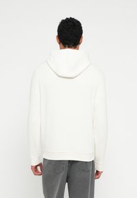 Biała bluza z kapturem, cechująca się miękką fakturą, długimi rękawami oraz prążkowanym wykończeniem na dole. Noszona z szarymi dżinsami, widziana od tyłu.