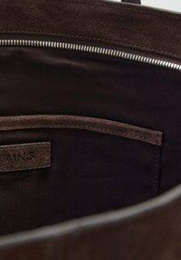 Wnętrze brązowej torebki zamszowej z gładką podszewką, z zamknięciem na zamek błyskawiczny, kieszonką oraz metką wytłoczoną napisem "MNG."