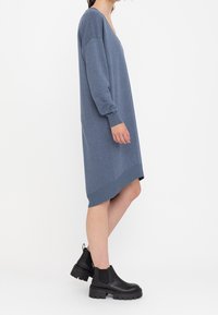 Abito maglione blu a V con maniche lunghe, polsini e orlo a costine, che arriva sopra il ginocchio. Abbinato a stivali neri alla caviglia.