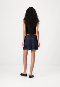 Jupe en denim bleu foncé avec un ourlet en volants, taille haute et poches arrière. Associée à un haut sans manches noir et des ballerines noires.