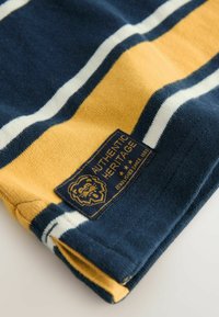 Kötött pulóver tengerészkék, sárga és fehér csíkokkal; tartalmaz egy anyagcímkét "AUTHENTIC HERITAGE" felirattal és egy oroszlán logóval.