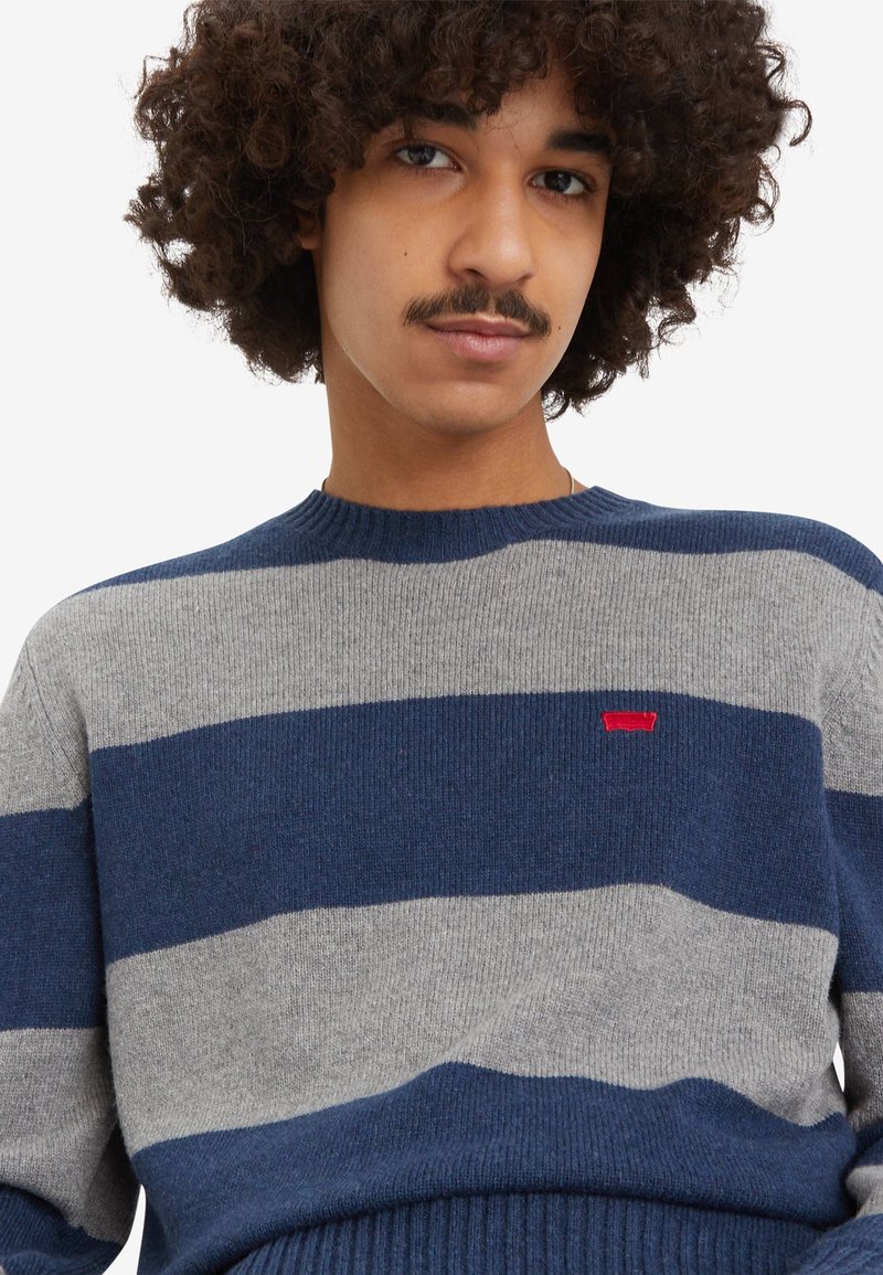 Suéter a rayas en mezcla de lana en colores azul marino y gris, con un cuello y dobladillo de canalé, y un pequeño logo rojo en el pecho.