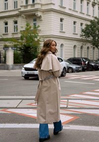Žena s dlouhými vlasy, oblečená v béžovém trenčkotě a modrých džínech, kráčí poblíž zaparkovaných aut na městské ulici, v pozadí je světlá budova.