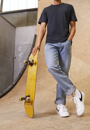 T-shirt nera a maniche corte, jeans in denim azzurro chiaro, skateboard giallo con ruote nere e sneakers bianche con dettagli neri.