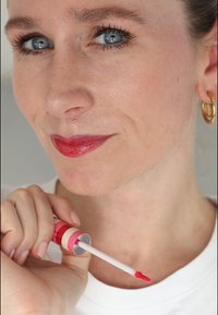 Läppfärg i en randig rosa och vit behållare, med en applikator i form av en spetssvans. Modellen visar en livfull röd läppfärg och en neutral hudton.