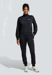 Γυναίκα που στέκεται φορώντας μαύρο tracksuit της Adidas με λευκά sneakers της Adidas, με τα χέρια χαλαρά, χαμογελώντας, μπροστά σε απλό ανοιχτό γκρι φόντο.