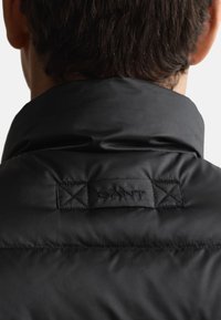 Schwarze Steppjacke mit hohem Kragen, aus strukturiertem Stoff, mit einem geprägten Logo-Patch und Kreuzstich-Detail an der Rückseite.