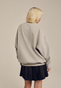 Túlméretes, világosszürke pulóver bordás mandzsettával, fekete pliszírozott szoknyával párosítva, hátulról nézve, semleges háttér előtt.