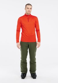 Punainen pitkähihaisessa urheilupaidassa, jossa on vetoketjukaulus, yhdistetty vihreisiin housuihin ja mustiin hiihtokenkiin ulkoilma-aktiviteetteihin. Silkkinen rakenne, istuva muotoilu.