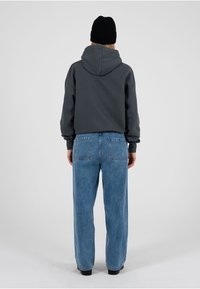 Grauer Kapuzenpullover mit lockerem Schnitt, kombiniert mit hellblauen, hoch taillierten Jeans mit zwei Gesäßtaschen. Schwarze Strickmütze auf dem Kopf.