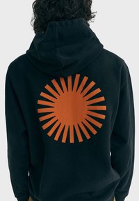 Čierna mikina s kapucňou s veľkým oranžovým slnečným lúčom na chrbte. Materiál pôsobí mäkko, s voľným strihom a kapucňou so šnúrkou na stiahnutie.