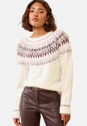 Junge Frau mit schulterlangem welligem Haar trägt einen weißen flauschigen Pullover mit lila Mustern und braune Lederhose.