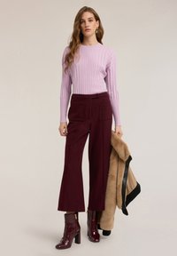 Top a maniche lunghe color lilla chiaro a coste, pantaloni ampi color bordeaux a gamba larga, stivaletti bordeaux con tacco alla caviglia. La modella tiene in mano un cappotto in pelliccia sintetica color cammello.