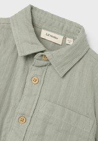 Camicia a maniche corte in cotone verde chiaro con colletto, bottoni in legno, texture a righe verticali e taschino sul petto.