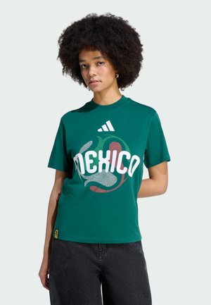 Kobieta o kręconych włosach w zielonej koszulce z napisem „Mexico” i logo Adidas, w czarnych spodniach, stoi na jednolitym tle.