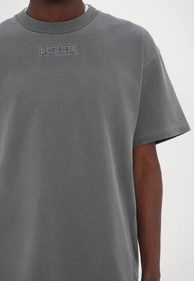 T-shirt en coton gris avec col rond côtelé, manches courtes et logo "KSUB" texturé au centre de la poitrine. Texture de tissu lisse.