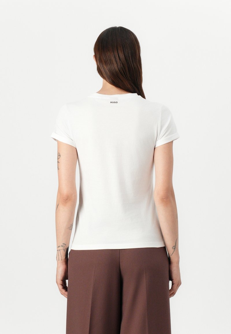 Femme aux longs cheveux bruns portant un t-shirt blanc manches courtes et un pantalon marron, debout de dos avec des tatouages visibles sur les bras.
