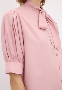 Blusa rosa con colletto annodato, maniche corte a sbuffo e chiusura frontale con bottoni. Realizzata in tessuto liscio con una sottile trama.